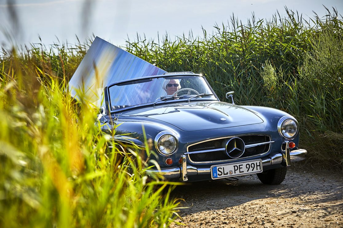 Kuenstlerin faehrt im Oldtimer mit Leinwand zum Strand